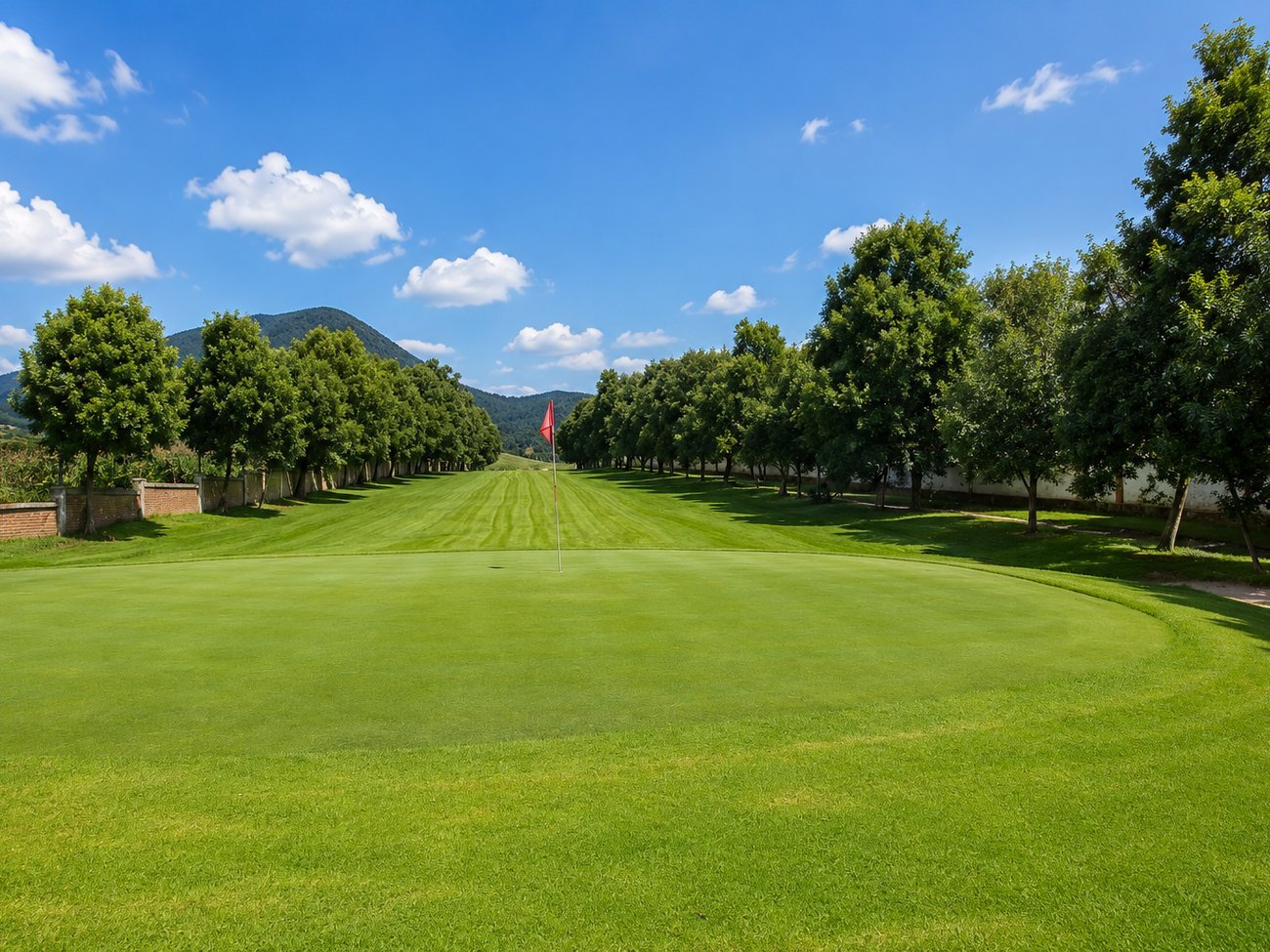 Green del campo de golf con banderín rojo entre árboles y vista a la montaña